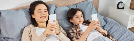 Eine Frau und ein Mädchen liegen auf einem Bett, konzentrieren sich auf ihre Mobiltelefone und genießen die gemeinsame Zeit zu Hause.