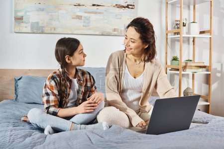 Téléchargez les photos : Une mère et sa fille assises sur un lit, englouties dans un ordinateur portable ensemble. - en image libre de droit
