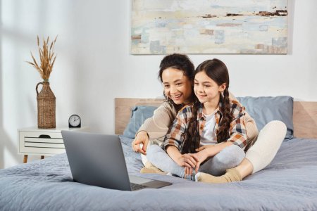 Foto de Madre e hija se sientan en una cama, absortos en la pantalla de un ordenador portátil, dedicándose a pasar tiempo juntos en casa. - Imagen libre de derechos