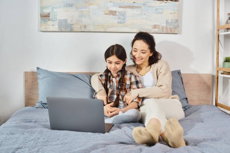 Foto de Una madre y una hija se sientan de cerca en una cama, enfocadas en una pantalla de computadora portátil con interés y conexión. - Imagen libre de derechos
