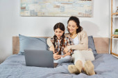 A mother and daughter sit closely on a bed, focused on a laptop screen with interest and connection. Stickers #698165874