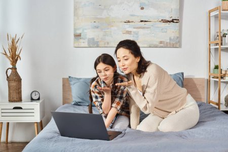 A woman and a girl share a tender moment on a bed while gazing at a laptop screen together.