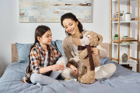 Una madre y una hija se sientan en una cama, disfrutando de un tiempo de calidad con un oso de peluche entre ellas.