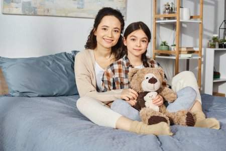 Téléchargez les photos : Deux femmes, une mère et sa fille, s'assoient sur un lit avec un ours en peluche, partageant un moment de proximité et de connexion. - en image libre de droit