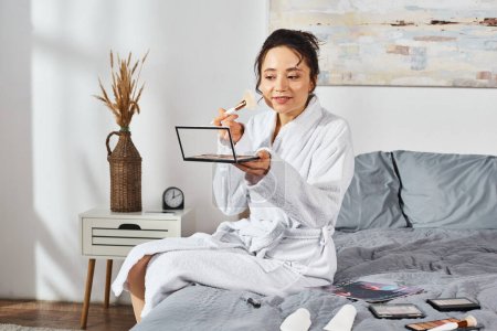 Eine brünette Frau im weißen Bademantel sitzt auf einem Bett mit Lidschatten, umgeben von Kosmetika und schminkt sich morgens..