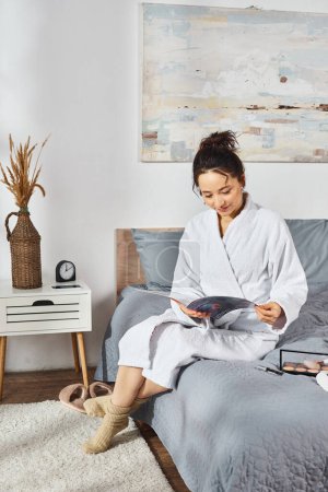 A brunette woman in a white bathrobe sits on a bed, engrossed in a magazine, with cosmetics scattered around her as she applies makeup.