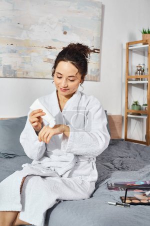 Una mujer morena sentada en una cama en un albornoz blanco, aplicando crema por la mañana.