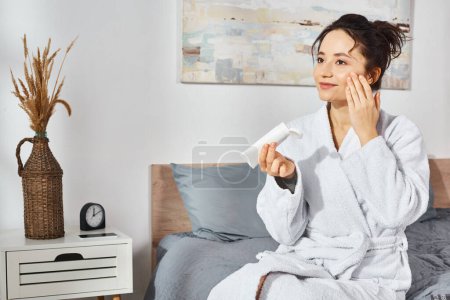 Eine brünette Frau im weißen Bademantel sitzt auf einem Bett mit Kosmetika und cremt sich morgens ein.