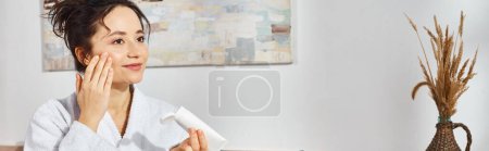 Téléchargez les photos : Une femme brune en peignoir blanc assise sur un lit avec des cosmétiques et appliquant de la crème visage - en image libre de droit