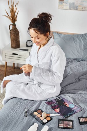 Une femme brune en peignoir blanc s'assoit sur un lit, concentrée sur sa crème tout en étant entourée de cosmétiques.