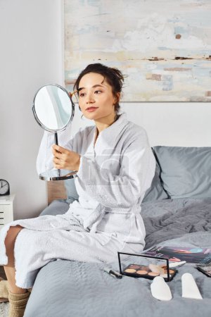 Une femme brune en peignoir blanc s'assoit sur un lit, examinant soigneusement quelque chose avec un miroir entouré de cosmétiques.