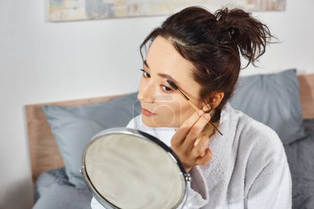 Téléchargez les photos : Une femme brune en robe blanche s'assoit sur un lit, tenant un miroir devant son visage et appliquant du maquillage le matin. - en image libre de droit