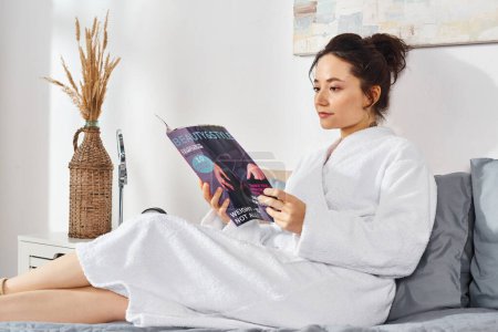 A brunette woman in a white bathrobe sits on a bed, immersed in reading a magazine, surrounded by cosmetics