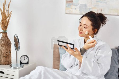 A brunette woman in a white bathrobe sits on a bed, apply makeup in the morning.