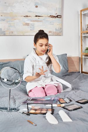 Foto de Una niña preadolescente en un albornoz blanco se sienta en una cama charlando en su teléfono celular. - Imagen libre de derechos