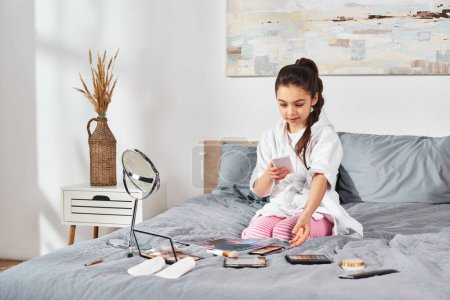 Téléchargez les photos : Une jeune fille brune en peignoir blanc s'assoit sur un lit, absorbée dans son téléphone portable. - en image libre de droit