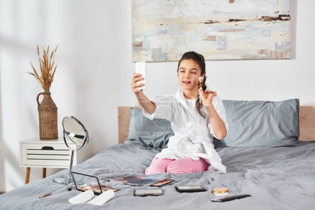 Foto de Una chica preadolescente morena en un albornoz blanco se sienta en una cama, sosteniendo un teléfono celular. - Imagen libre de derechos