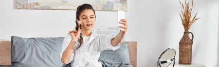 Téléchargez les photos : Une préadolescente brune en peignoir blanc s'assoit sur un lit tenant un téléphone portable. - en image libre de droit