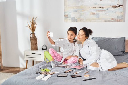 Téléchargez les photos : Mère et fille en peignoirs blancs s'assoient sereinement ensemble sur le lit, partageant un moment paisible de liaison. - en image libre de droit