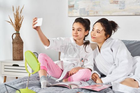 Téléchargez les photos : Une mère brune et sa fille en peignoirs blancs confortablement assises sur un lit, partageant un moment de paix et de convivialité. - en image libre de droit