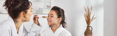 Photo for A brunette mother in a white bathrobe is delicately applying face powder on her daughters face while sitting on a bed. - Royalty Free Image