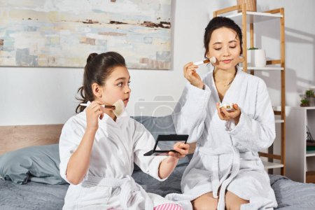 A brunette mother and daughter sit peacefully on a bed, wrapped in white bath robes, applying makeup