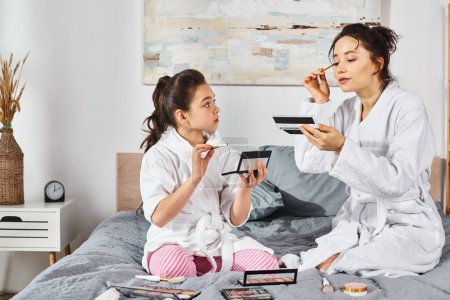 Photo for A brunette mother and daughter in white bath robes sit on a bed applying makeup together. - Royalty Free Image
