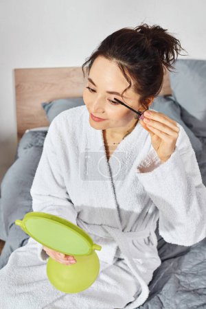 Una mujer con cabello moreno se aplica rímel en las pestañas mientras está sentada en una cama