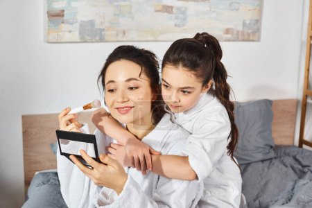 Téléchargez les photos : Une mère brune et sa fille en peignoirs blancs s'assoient ensemble sur un lit confortable, avec une palette de fards à paupières - en image libre de droit