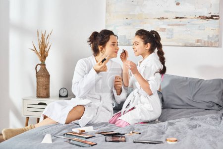 Photo for A brunette mother and daughter in white bath robes sit peacefully on a bed, sharing a tender moment together. - Royalty Free Image