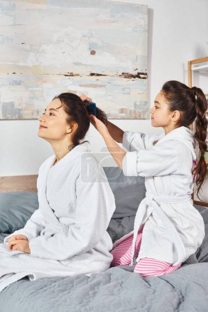 A brunette mother and daughter, both in white bath robes, sit peacefully on a cozy bed.