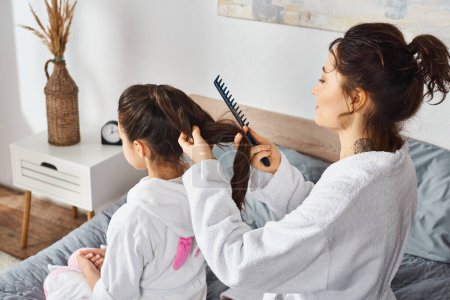 Photo for A brunette mother in a white bathrobe sits on a bed combing the hair of her young daughter, also in a white bathrobe. - Royalty Free Image