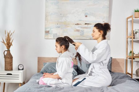 Téléchargez les photos : Brunette mère et fille en peignoirs blancs assis sur le lit, partageant un moment d'amour pendant que la mère brosse les cheveux des filles. - en image libre de droit