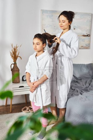 Téléchargez les photos : Une mère brune et sa fille en peignoirs blancs se tenant ensemble dans une chambre, partageant un moment spécial. - en image libre de droit