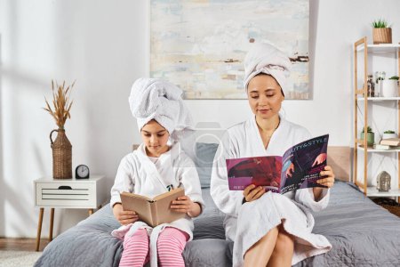 A brunette mother and daughter in white bath robes sit on a bed, immersed in a book and magazine