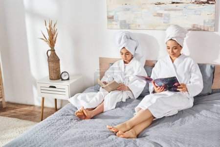 A brunette mother and daughter in white bath robes sit together on a bed, reading a book and enjoying each others company.