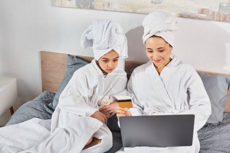 Two women, a brunette mother, and daughter, in white bath robes sitting on a bed, engaged with a laptop.