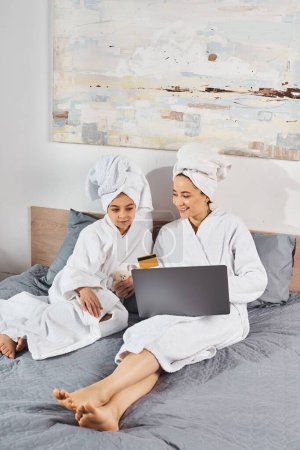 A brunette mother and daughter in white bath robes peacefully sitting together on top of a bed.
