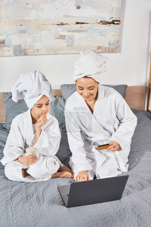 Photo for A brunette mother and daughter are sitting together on top of a bed, both wearing white bath robes. - Royalty Free Image
