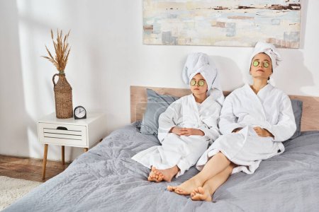 Eine brünette Mutter und ihre Tochter sitzen in weißen Bademänteln auf einem Bett, Handtücher um den Kopf gewickelt.