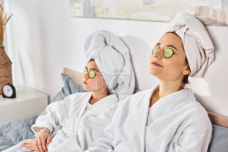 Zwei brünette Frauen in weißen Bademänteln genießen eine Wellness-Behandlung mit Gurkenpflaster auf ihren Augen.