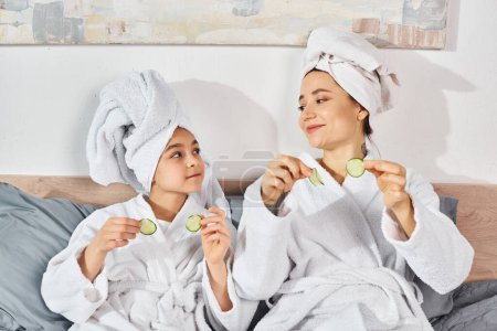 Brünette Mutter und Tochter genießen einen friedlichen Moment, während sie in passenden weißen Roben auf einem Bett sitzen, Gurken in der Hand, Schönheit