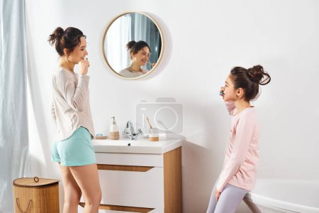 A brunette woman and her preteen daughter stand together in front of a bathroom mirror, engaging in their beauty and hygiene routine.