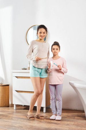 A brunette woman stands with her preteen daughter in a modern bathroom, engaging in beauty and hygiene routines together.