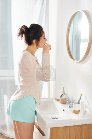 A brunette woman brushes her teeth in front of a bathroom mirror in morning time
