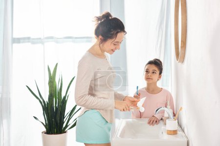 A brunette woman and her preteen daughter are standing next to each other in a modern bathroom sink, engaged in a beauty and hygiene routine.