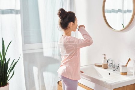 A brunette woman in a modern bathroom is brushing her teeth as part of her beauty and hygiene routine.
