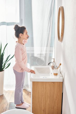 A brunette girl stand in a modern bathroom, brushing teeth in front of a mirror.