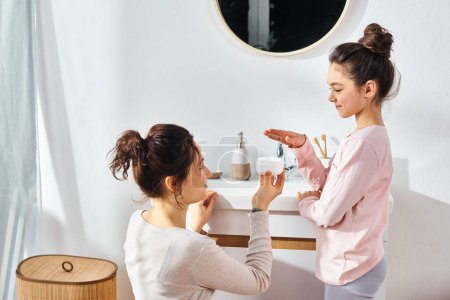 A brunette woman and her preteen daughter are standing together in a modern bathroom, engaged in their beauty and hygiene routine.