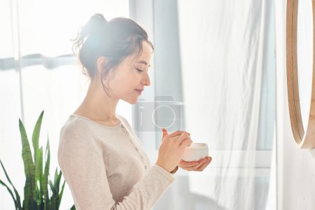 Una mujer morena se para frente a un espejo sosteniendo un frasco de crema, dedicándose a su rutina de belleza e higiene matutina.